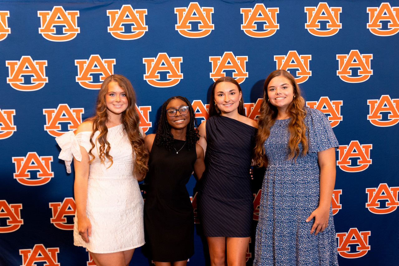 A group of summer program participants smiling for the camera in front of an Auburn University step and repeat using the university's logo.