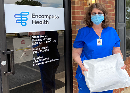 Woman with glasses and wearing scrubs and a mask stands in front of Encompass Health with envelope full of donated face masks.