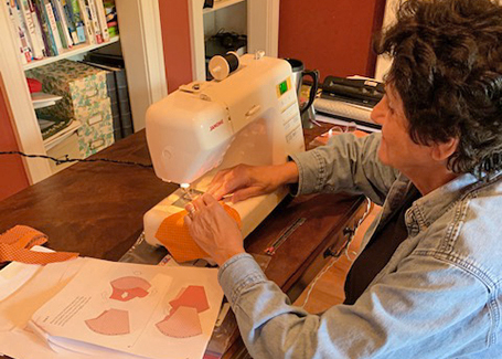 Carol Womer sitting at sewing machine with back to camera sewing a fabric face mask.