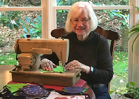 Older woman sits behind sewing machine with windows behind her.
