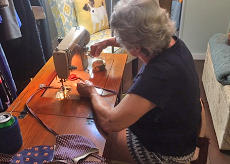 Doris Miller sits behind sewing machine sewing orange and blue masks.