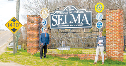 Two men stand on either side of the 'Welcome to Historic Selma' town sign