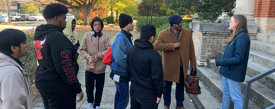 A group of male and female students stand outside for a tour while listening to a female tour guide