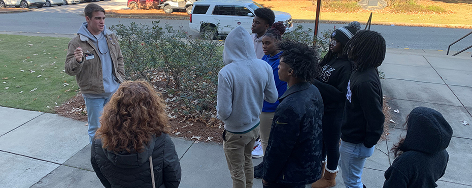 A group of male and female students stand outside for a tour while listening to a male tour guide