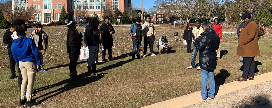 A group of male and female students stand outside in a crop field to learn more from a speaker about agricultural production