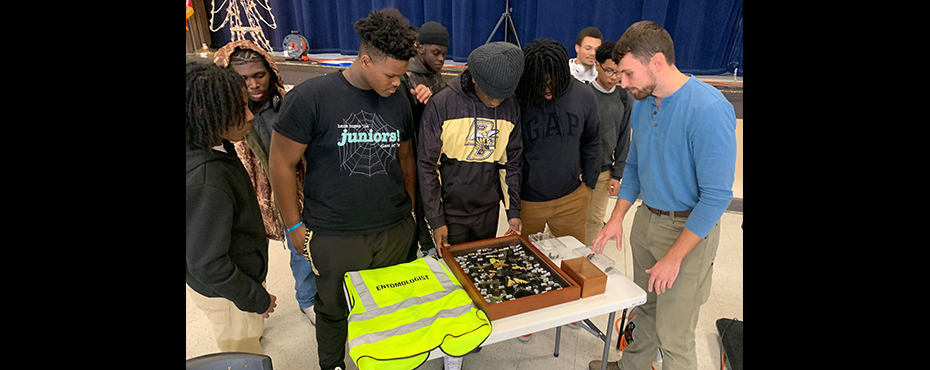 A small group of students observe an insect collection display with a male guest speaker explaining