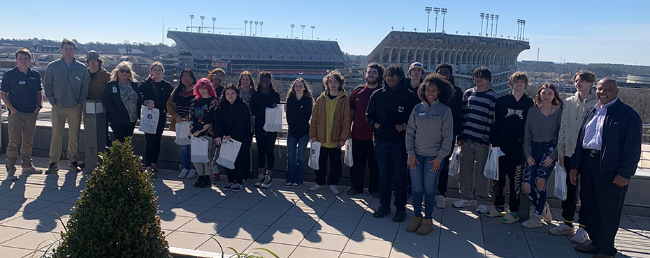    A large group of male and female tour guides, students, and teachers stand on a terrace for a picture with a large football stadium in the background