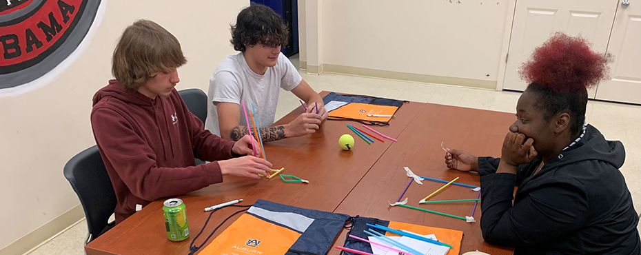 Two male students and one female student sit at a table working on a project with straws, a tennis ball and tape.