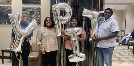 Four students standing and holding individual silver balloons with the letters YPIT for Young Professionals In Training