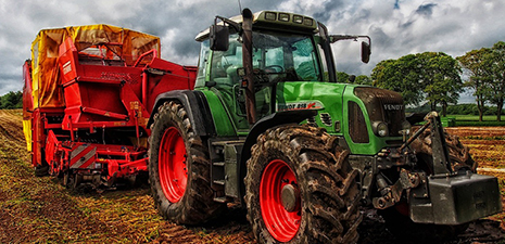 Green tractor pulling a farming implement in a field surrounded by trees to the right and blue sky and clouds