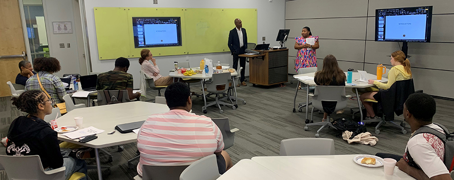    A male speaker presents to a group of male and female students in a modern classroom environment.