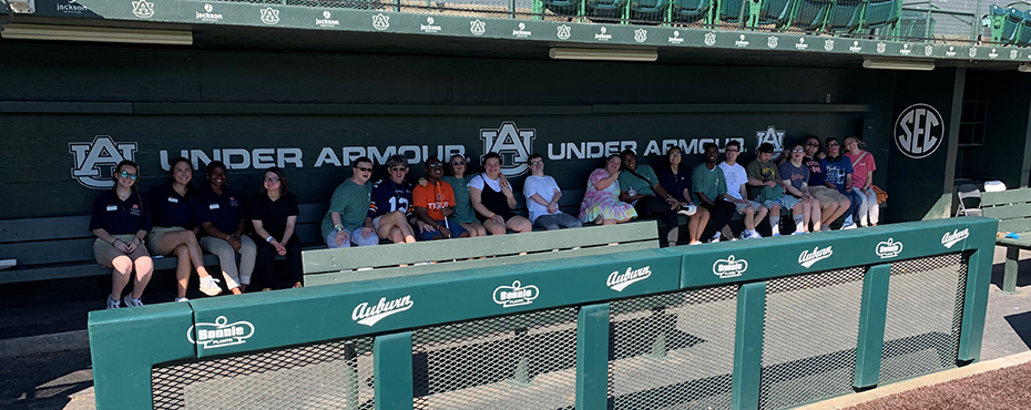 A large group of male and female students and adults sit in the 1st base dugout for a group picture