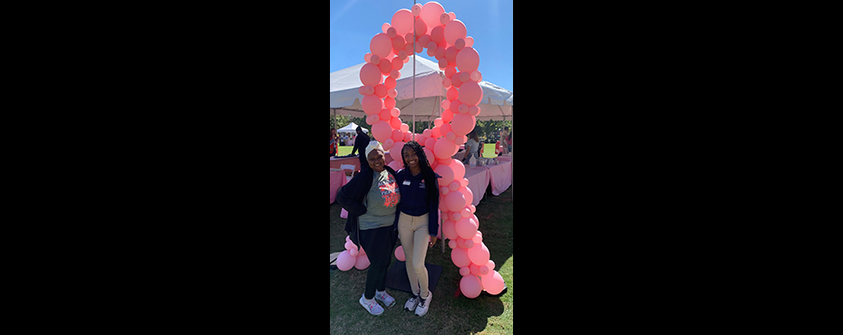 2 female students stand and smile for a picture at an outreach event with pink balloons backdrop