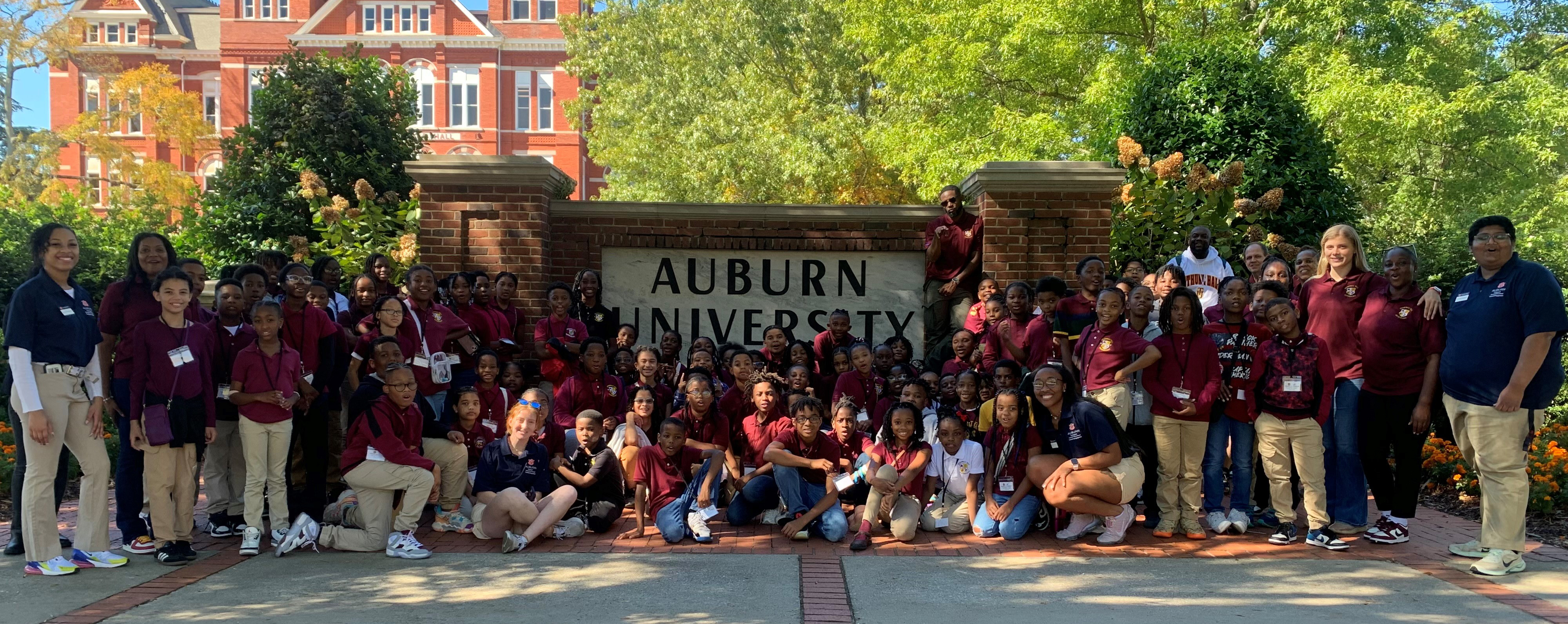 A large group of male and female students, teachers, and outreach ambassadors stand for a group picture in front of the Auburn University sign and Samford Hall backdrop