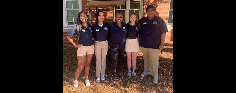 3 female students, 1 male student, and 1 female adult stand for a picture under a shade tree outside on campus