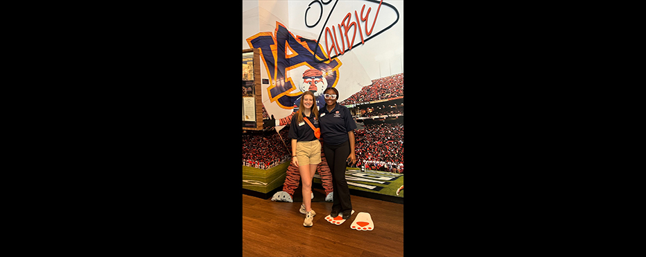 2 female students leading a tour smile for a picture with an Auburn flag and football field backdrop.