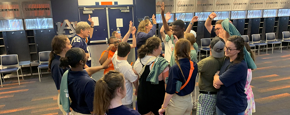 A large group of male and female students and adults stand in a game room and cheer on after a huddle