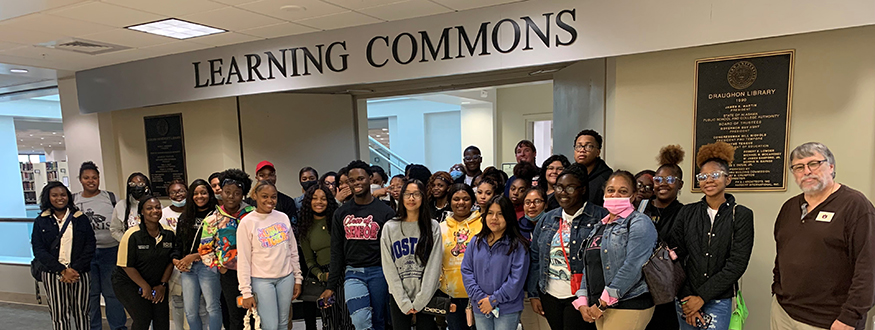 A large group of male and female students and adults smile at the conclusion of a library tour and visit under a large transition sign that reads Learning Commons.