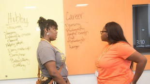 Two young black girls talk to each other in front of dry erase board.