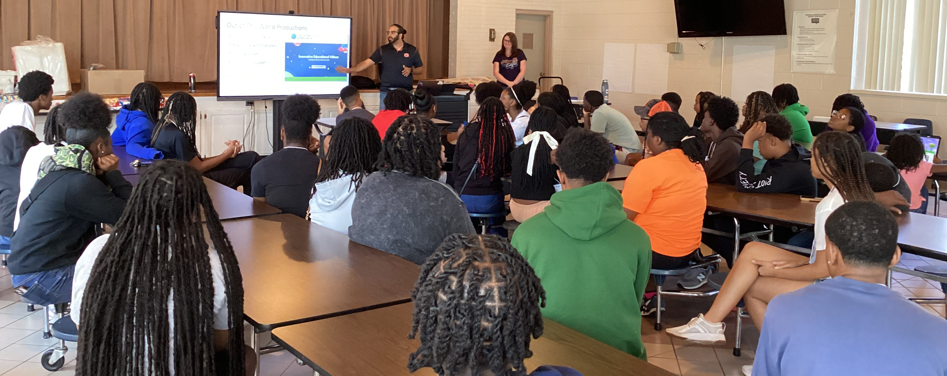 A male guest speaker stands and presents to a large group of male and female students in an auditorium