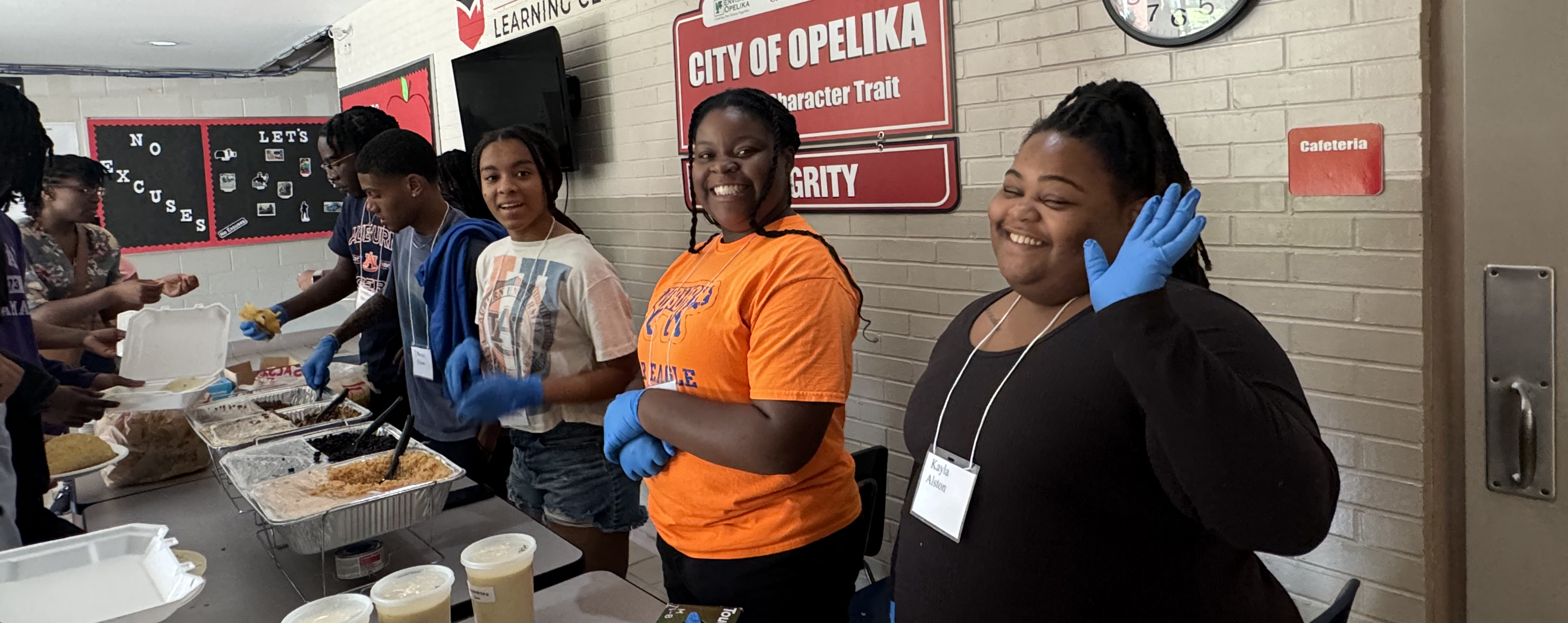 3 female students and 2 male students wave and smile for a picture as they serve food to community members