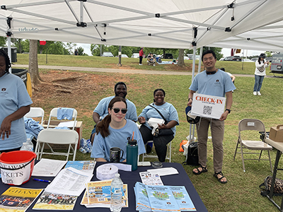 Several volunteers sitting at a table outside, one standing holding a check-in sign.