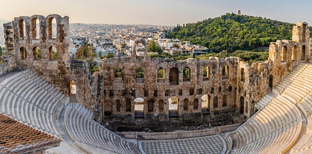 The sun shines on the ruins of an ancient Greek amphitheater with modern day Greece in the distance.