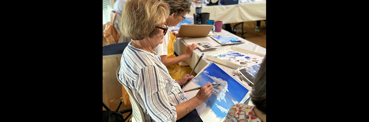 A woman adds fluffy white clouds to a sky blue canvas during an acrylic painting class.