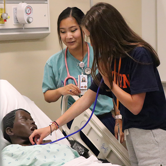 Two girls practicing taking blood pressure on a nursing patient mannequin in the bed