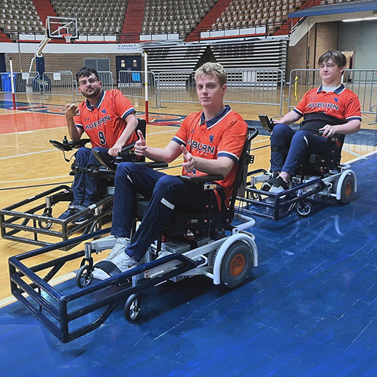 Three male students in powered wheelchairs on a basketball court