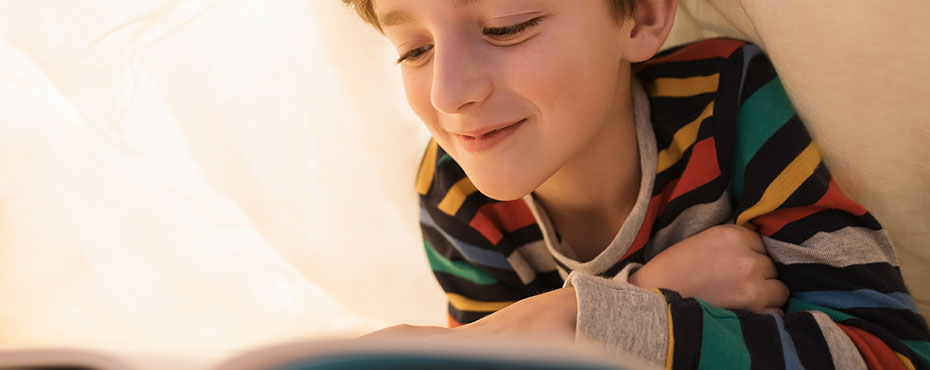 Young male under sheeting reading a book while wearing multi color striped shirt