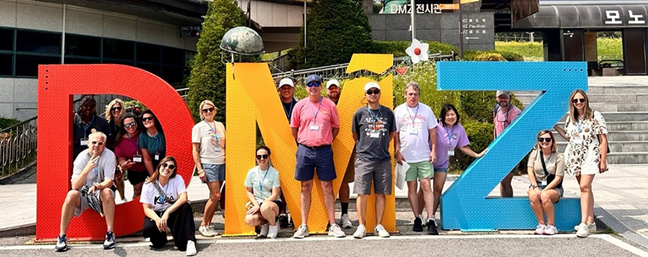 Group photo outside of mean and women standing around and in between 3 big red letters of DMZ.