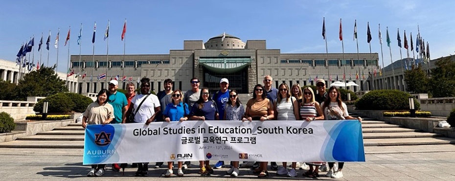 Group participants stand outside goverment building with multiple flags erected holding a large banner, Global Studies in Education South Korea.