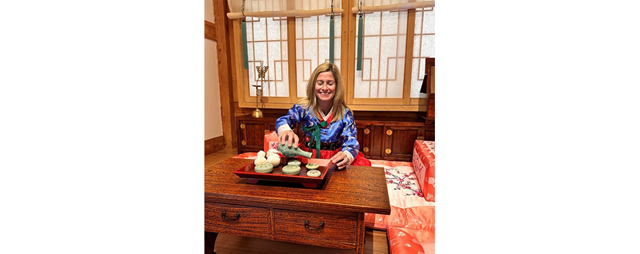 Woman dressed in traditional Korean dress sitting at table preparing tea.