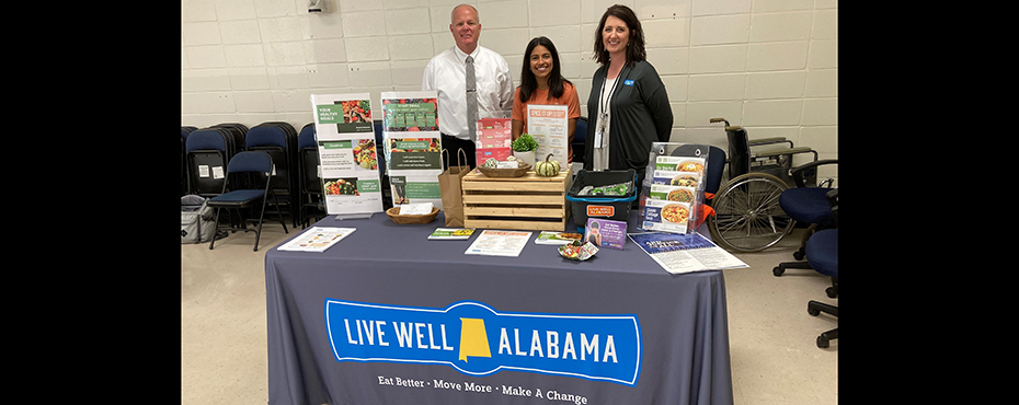 A man and two women pose for photo behind a table displaying promotional materials from Live Well Alabama - Eat better - Move More - Make a Change