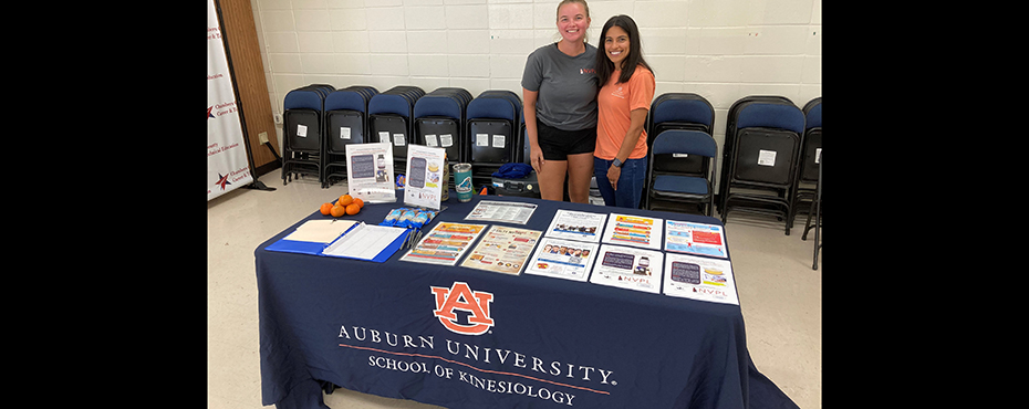 Two girls pose for photo behind a table displaying promotional materials from the AU School of Kinesiology